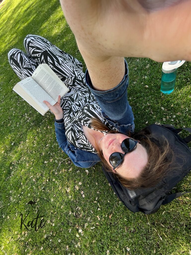 Kate laying on grass with her head on her backpack while she reads at the park