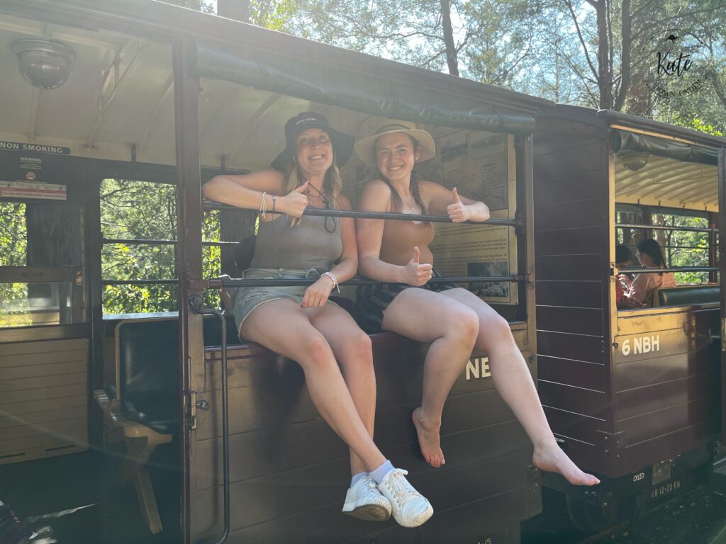 Kate and another woman in shorts sitting in a passenger rail car on the window sill, held in by horizontal bars, with their legs hanging outside the car