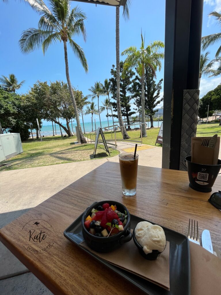 foreground: a bowl of fruit, dish of vanilla gelato, and iced latte on a light coloured wood table top
Background: palm trees and bright blue waters