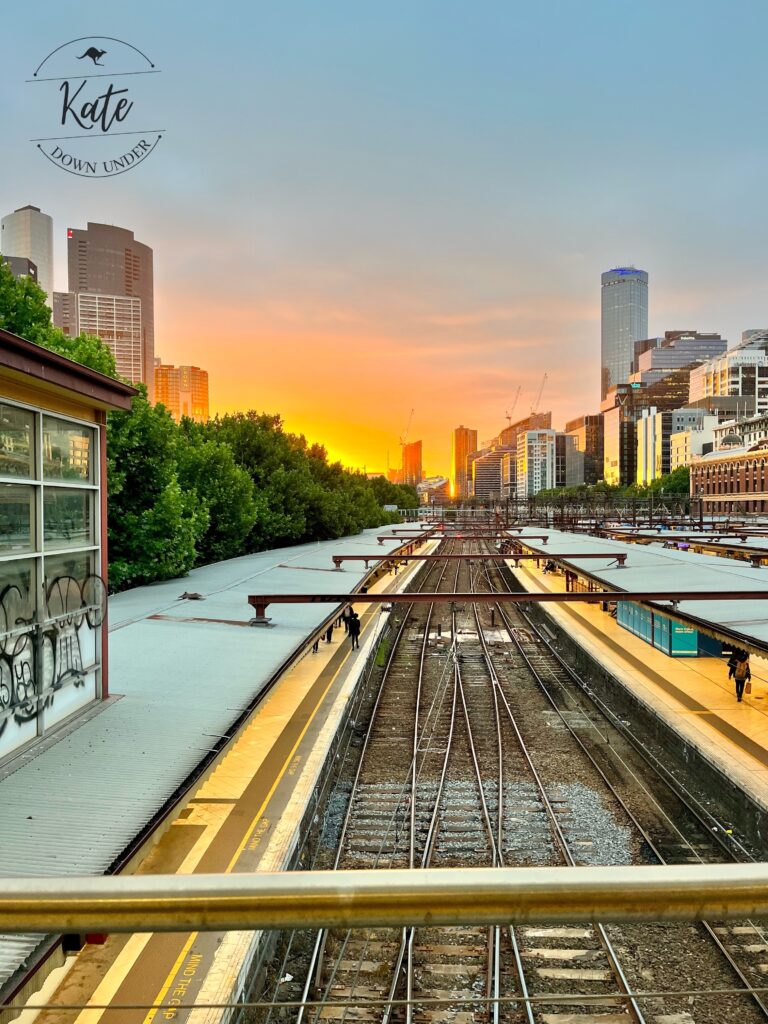 foreground of looking down onto the rail lines with platforms on either side at Flinders Street Station, with the sunset and city skyline in the background