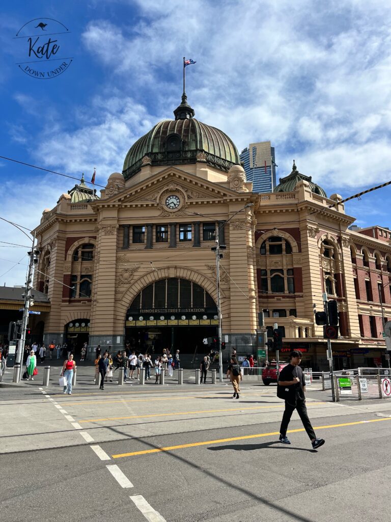 Flinders Street train station, with a partly cloudy background and several pedestrians crossing the street.