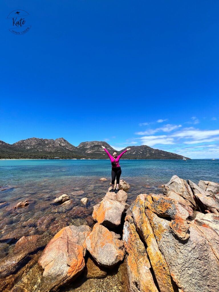 Kate standing on some partially submerged rocks with her hands in the air, in front of Coles Bay, in Freycinet National Park in Tasmania