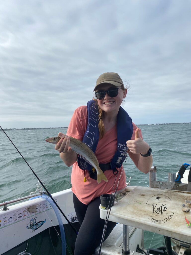 Kate standing on a boat holding the King George Whiting she caught with one hand around its gills, and giving a thumbs up with the other hand.