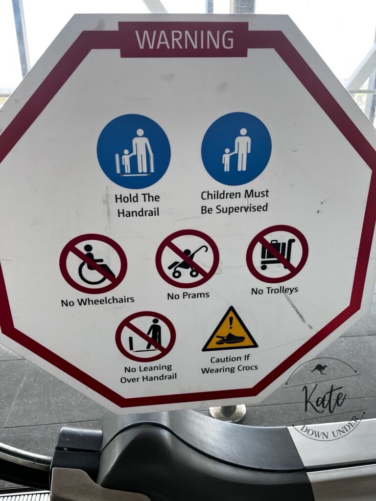A warning sign with various warnings about using the travelator with luggage, prams, wheelchairs, and a final disclaimer to use caution if wearing crocs